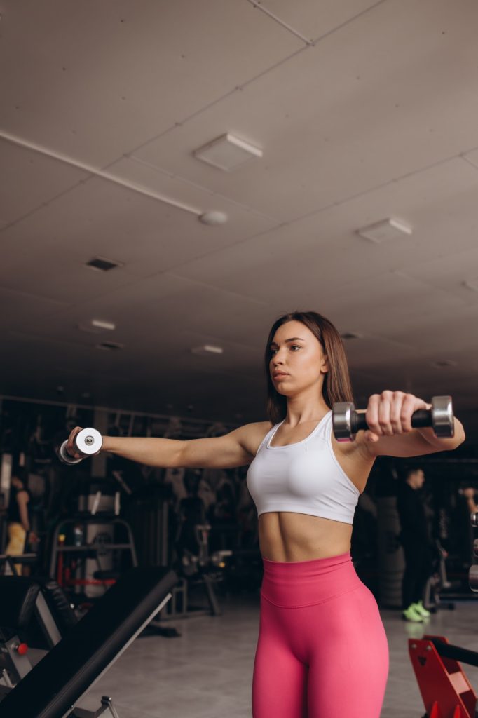 Attractive young fit sportswoman doing lateral raises smiling confidently working out with dumbbells