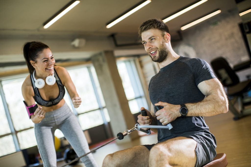 Young man using seated row machine in the gym with support of female coach