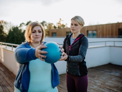 Overweight woman holding ball and exercising with personal trainer in outdoors on gym terrace