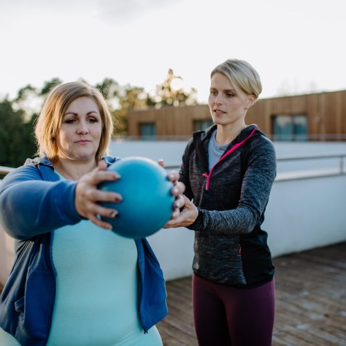 Overweight woman holding ball and exercising with personal trainer in outdoors on gym terrace